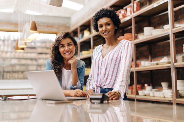 Cheerful female entrepreneurs smiling at the camera while working in their ceramic store. Happy young ceramists using a laptop together. Two young businesswomen running a successful small business.